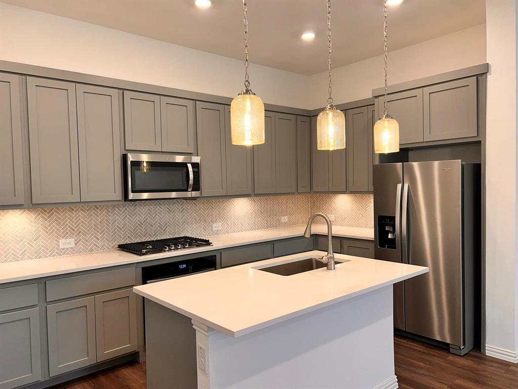 Kitchen featuring gray shaker cabinetry, stainless steel appliances, herringbone tile backsplash, an island with an integrated sink, and pendant lighting