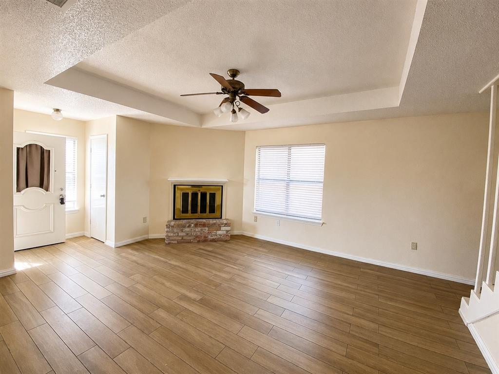 Spacious interior featuring wood-finish flooring, a tray ceiling with a ceiling fan, and a fireplace with a brick base Spacious interior featuring wood-finish flooring, a tray ceiling with a ceiling fan, and a fireplace with a brick base