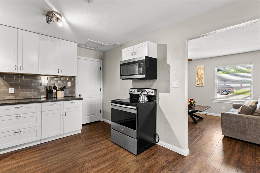 Kitchen featuring white cabinetry, dark countertops, a metallic subway tile backsplash, stainless steel appliances, and wood-finish flooring Kitchen featuring white cabinetry, dark countertops, a metallic subway tile backsplash, stainless steel appliances, and wood-finish flooring