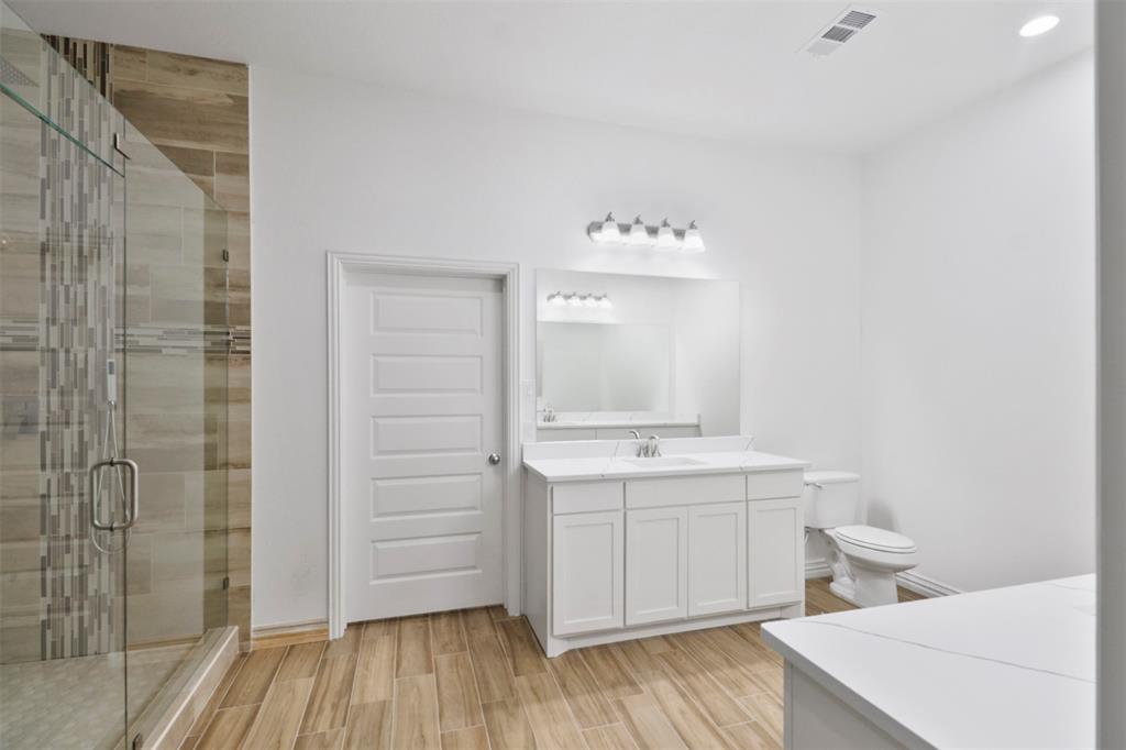 Spa-inspired primary bathroom featuring a frameless glass-enclosed shower with mixed-material tile surround, dual-sink vanity with white cabinetry and integrated lighting, wood-finish flooring, and a freestanding soaking tub