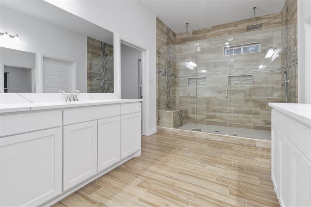 Spa-inspired primary bathroom featuring dual vanities with white cabinetry, a frameless glass-enclosed walk-in shower with tile surround and rainfall showerhead, wood-finish flooring throughout, and abundant natural lighting from transom windows
