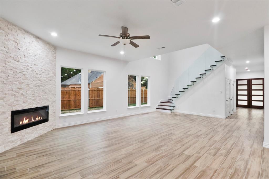Spacious living area featuring wood-finish flooring, a linear fireplace with a stacked stone surround, multiple windows, a ceiling fan, and a floating staircase with glass railings