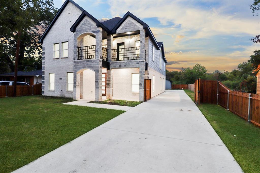 Contemporary exterior featuring white brick and stone accents, a dual second-story balcony with iron railings, a stone archway entryway, a concrete driveway, and a wooden privacy fence