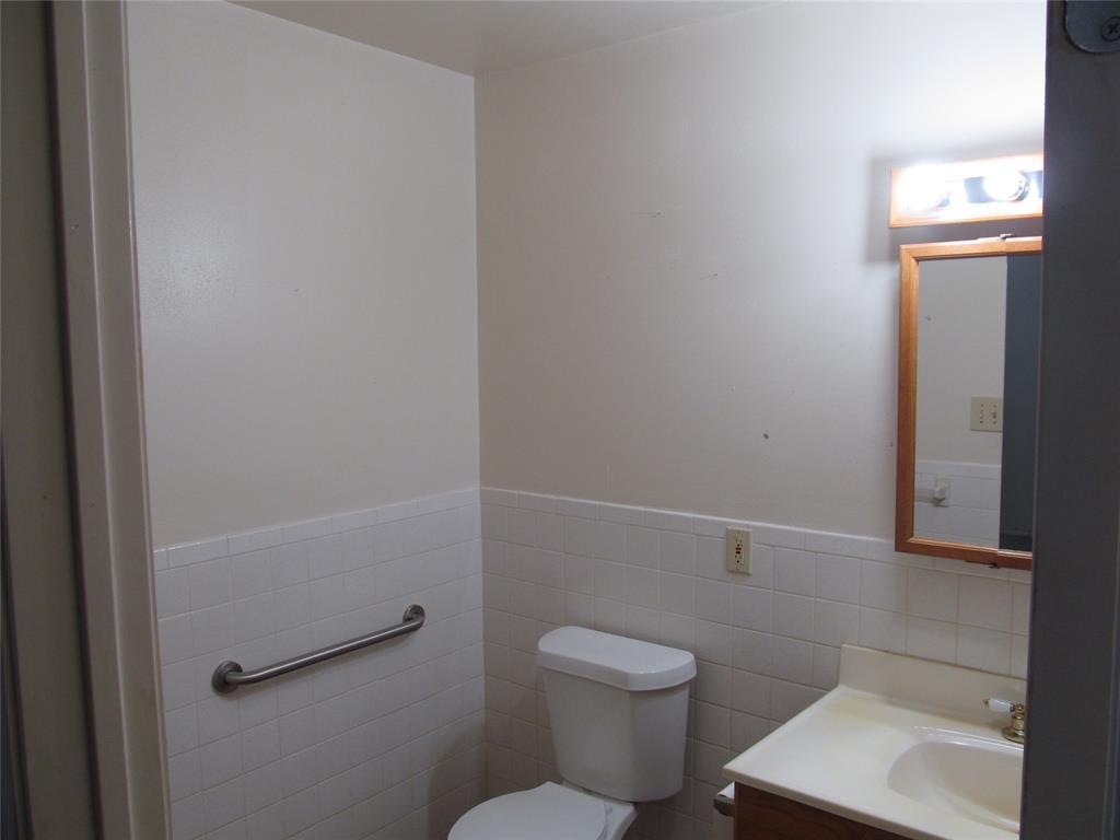 White tile wainscoting, built-in grab bar, wood-finish vanity with single sink, and wall-mounted mirror with integrated lighting