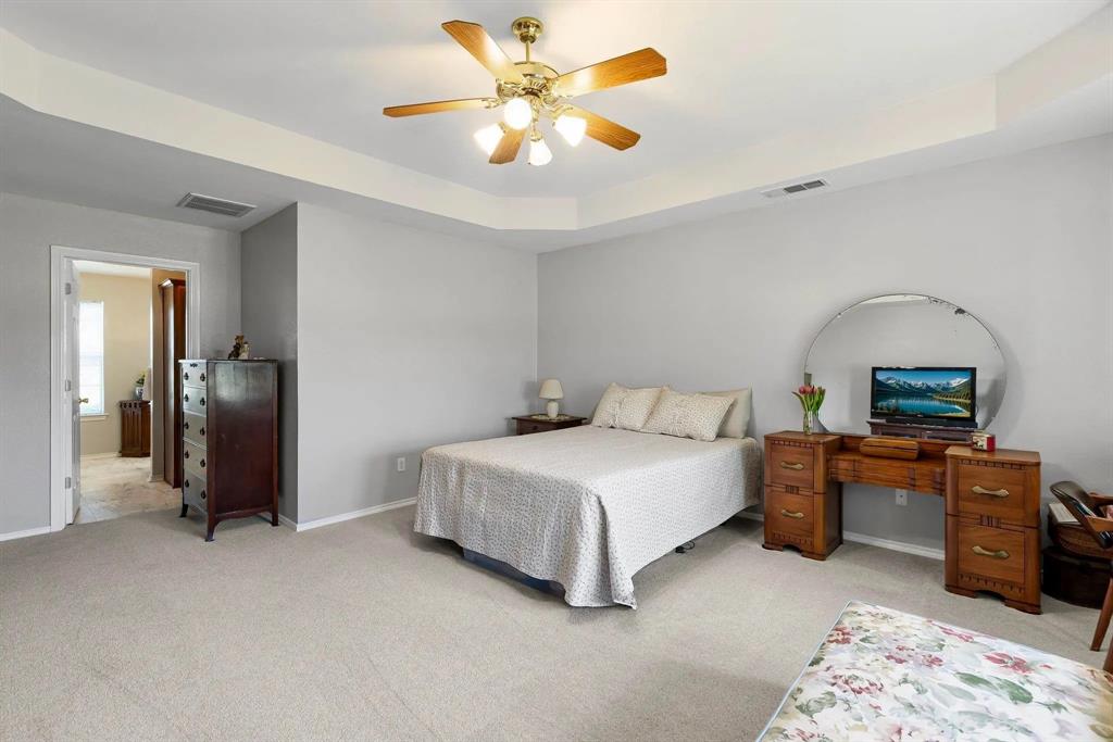Spacious bedroom featuring light grey walls, light-toned carpet flooring, and a tray ceiling with a ceiling fan