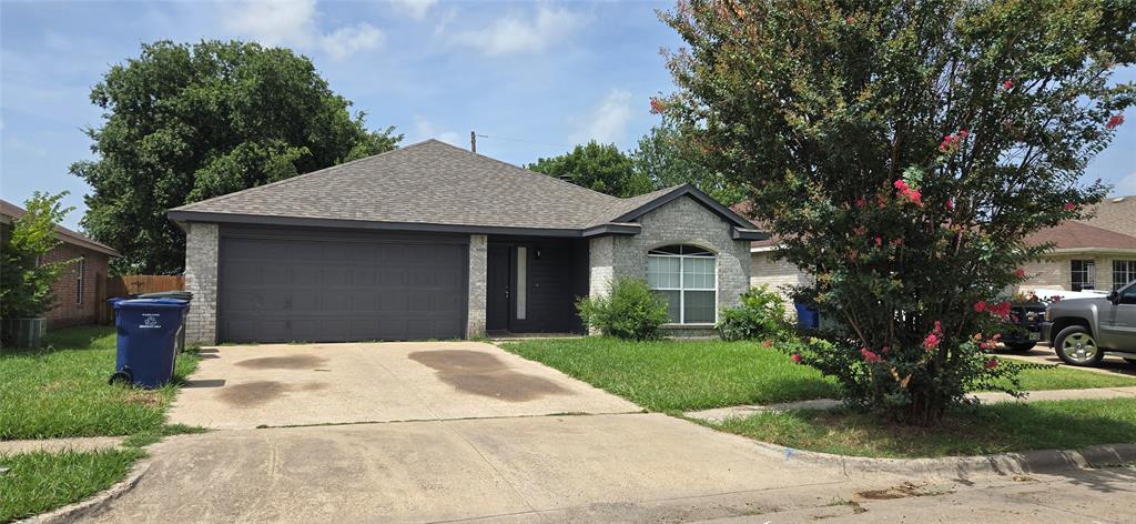 Gray brick exterior with a dark gray garage door, a gray shingle roof, a front lawn, and a mature tree with pink blooms Gray brick exterior with a dark gray garage door, a gray shingle roof, a front lawn, and a mature tree with pink blooms