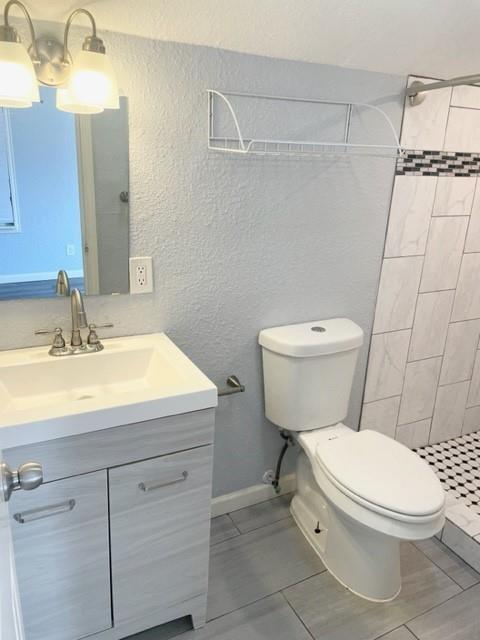 Bathroom featuring a single vanity with a white countertop and light wood-finish cabinetry, a commode, a shower with white and black mosaic tile flooring, and gray rectangular wall tile with a decorative accent band