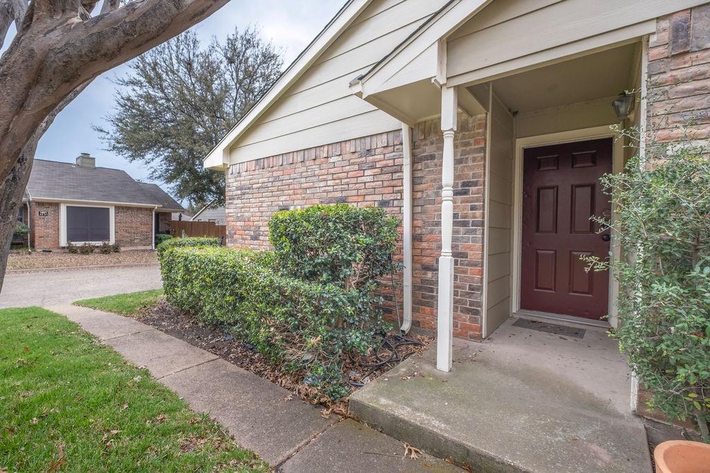 Brick and siding exterior featuring a covered entryway with a paneled front door, an established shrub border, and a concrete walkway Brick and siding exterior featuring a covered entryway with a paneled front door, an established shrub border, and a concrete walkway