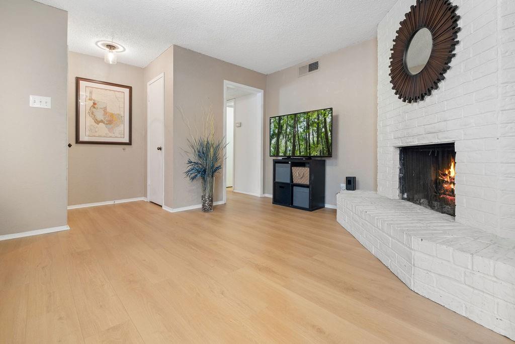 Expansive living area featuring light wood-finish flooring, a white-painted brick fireplace with a hearth, and neutral wall tones Expansive living area featuring light wood-finish flooring, a white-painted brick fireplace with a hearth, and neutral wall tones