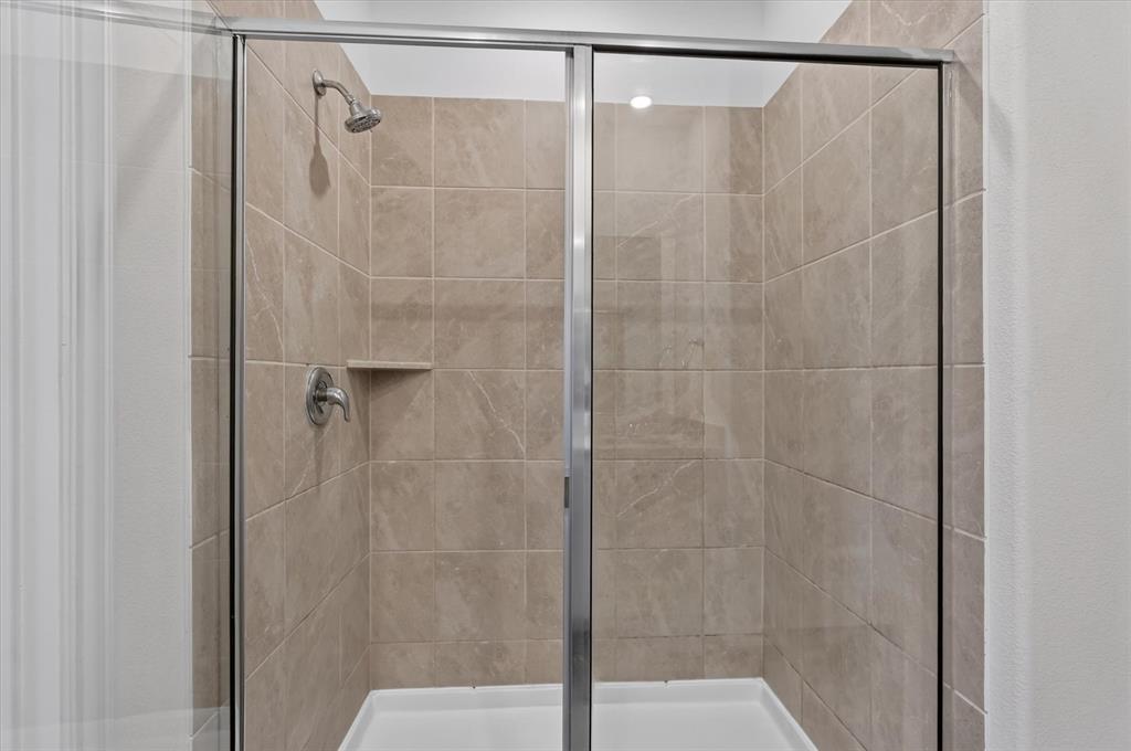 Spa-inspired shower enclosure featuring frameless glass doors with polished chrome hardware, neutral tile surround with linear layout, dual showerheads with individual controls, and a spacious walk-in configuration with built-in shelf niche