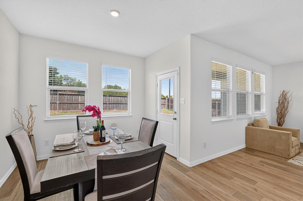Bright dining area featuring a wood-finish table with upholstered seating, abundant natural light from dual windows, and direct access to an adjacent room through a white entry door