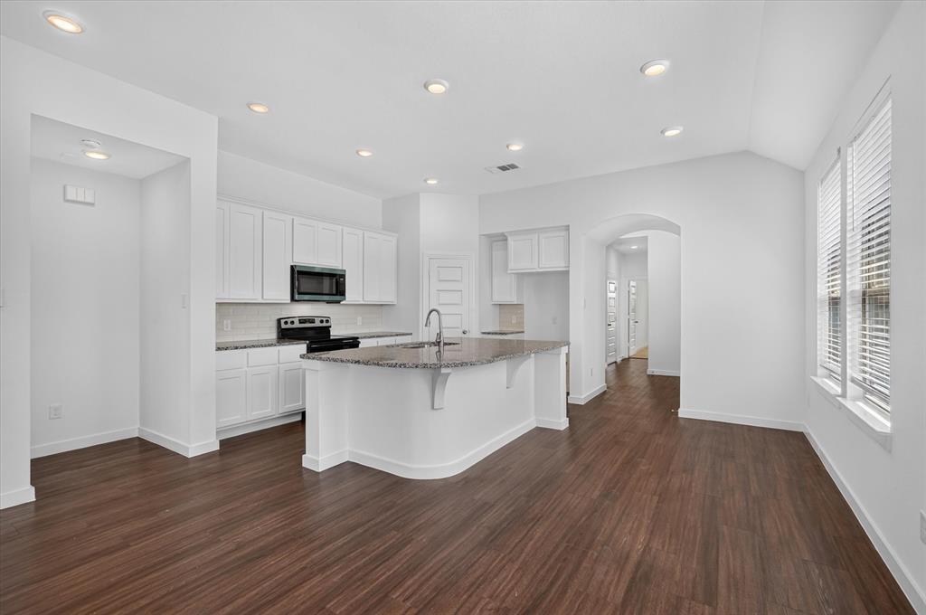 Modern kitchen and dining area featuring white cabinetry with stainless steel appliances, a curved island with dark countertop and undermount sink, recessed ceiling lighting, wood-finish flooring throughout, and an arched passageway leading to additional living spaces