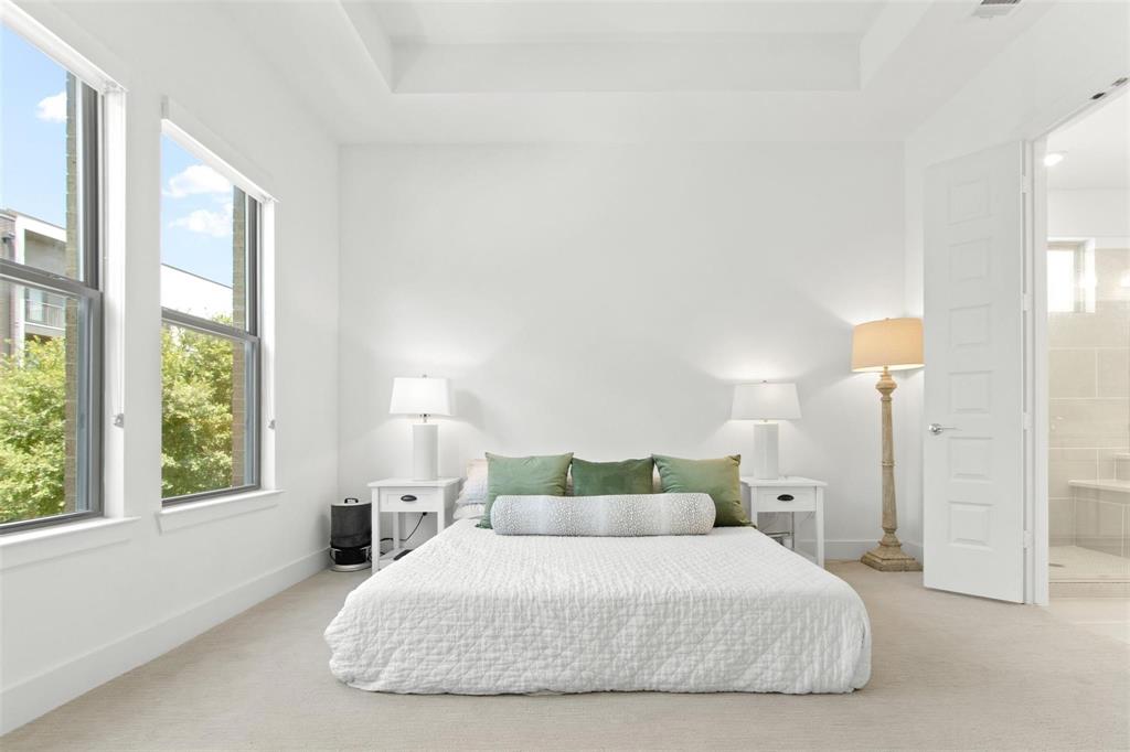 Carpeted room featuring a tray ceiling, two tall windows, and a panel door leading to a tiled bath area with a built-in bench