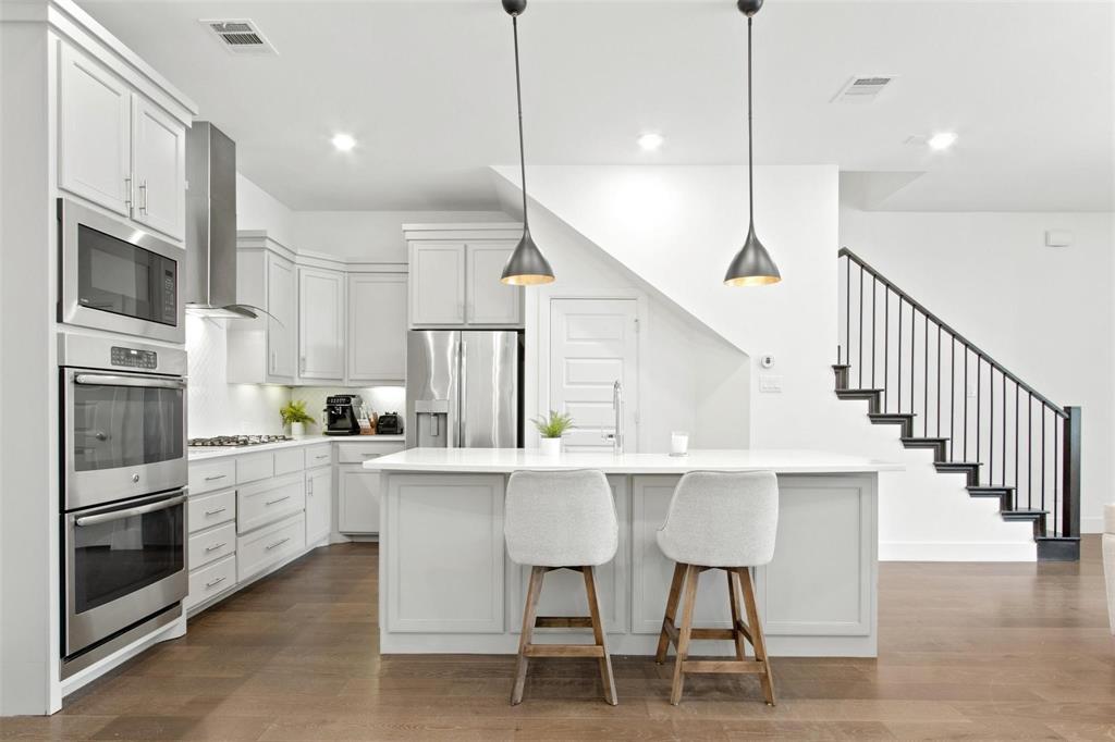 Modern kitchen featuring light cabinetry, stainless steel appliances, a center island with pendant lighting, wood-finish flooring, and a contemporary staircase with dark metal balusters