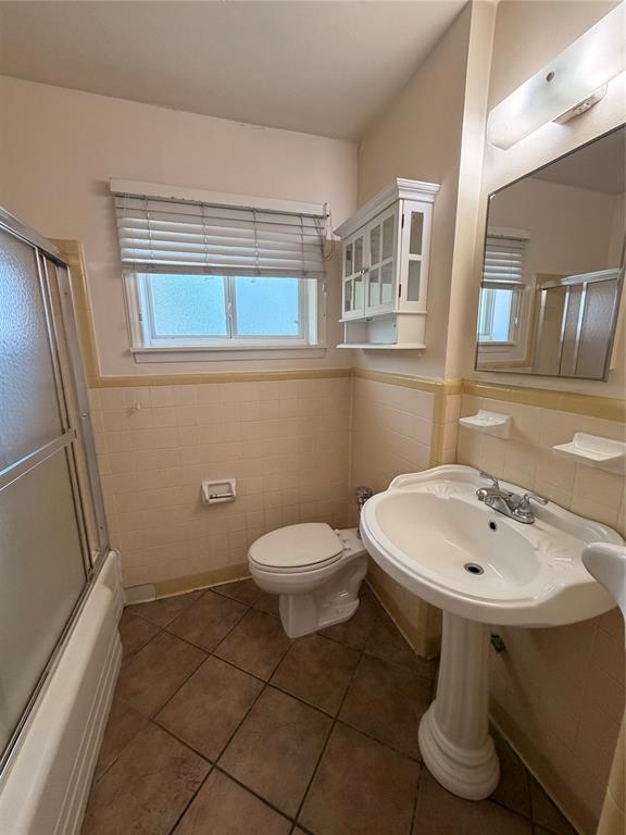 Bathroom featuring a ceramic pedestal sink, tiled wall wainscoting, glass-enclosed shower, wall-mounted white cabinetry, and ceramic tile flooring