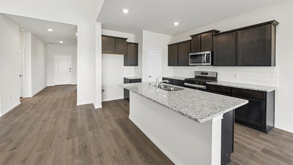 Kitchen featuring a granite island, dark wood-finish cabinetry, stainless steel appliances, a subway tile backsplash, and wood-finish flooring Kitchen featuring a granite island, dark wood-finish cabinetry, stainless steel appliances, a subway tile backsplash, and wood-finish flooring
