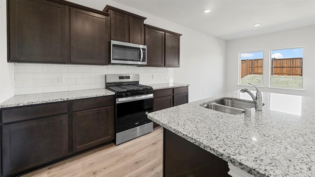 Modern kitchen featuring dark cabinetry, stainless steel gas range and microwave, granite-style countertops with waterfall edge, stainless steel single-basin sink with chrome faucet, subway tile backsplash, and wood-finish flooring throughout Modern kitchen featuring dark cabinetry, stainless steel gas range and microwave, granite-style countertops with waterfall edge, stainless steel single-basin sink with chrome faucet, subway tile backsplash, and wood-finish flooring throughout