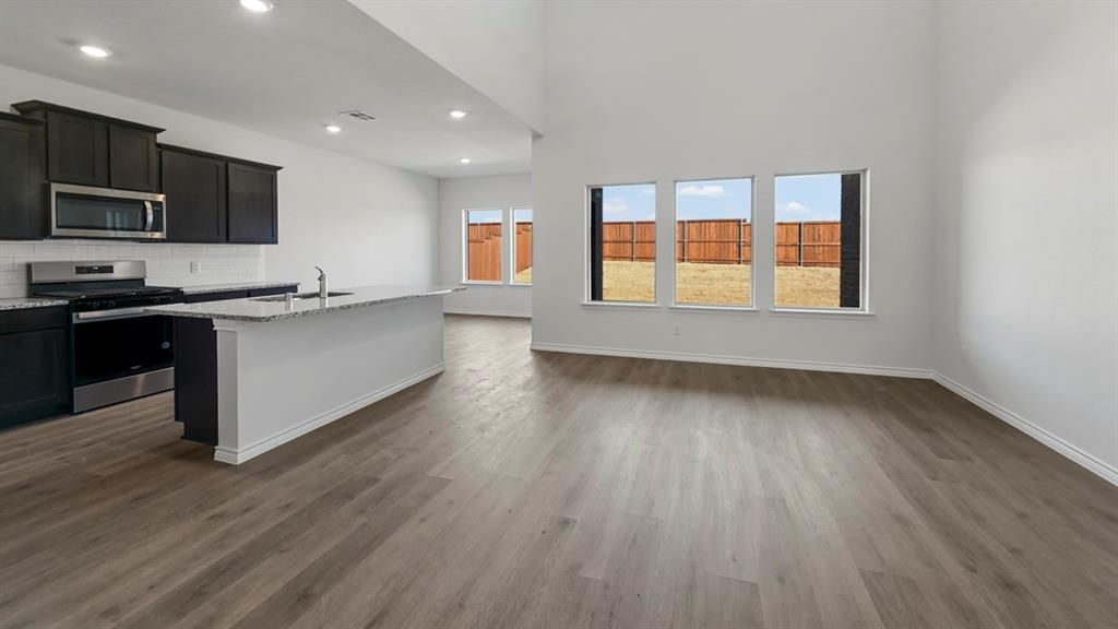 Spacious common area featuring wood-finish flooring, a kitchen island with sink, dark cabinetry, stainless steel appliances, and multiple windows