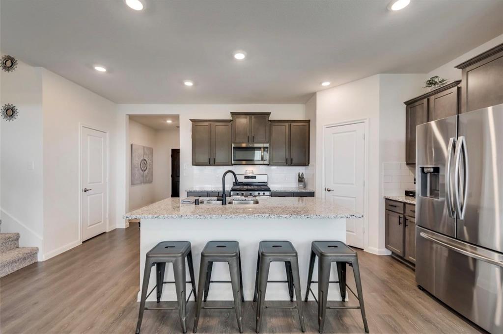 Modern kitchen featuring a spacious white quartz island with seating for four, stainless steel appliances including a gas cooktop and side-by-side refrigerator, dark cabinetry with integrated hardware, recessed ceiling lighting, wood-finish flooring, and an open layout connecting to the adjoining living spaces