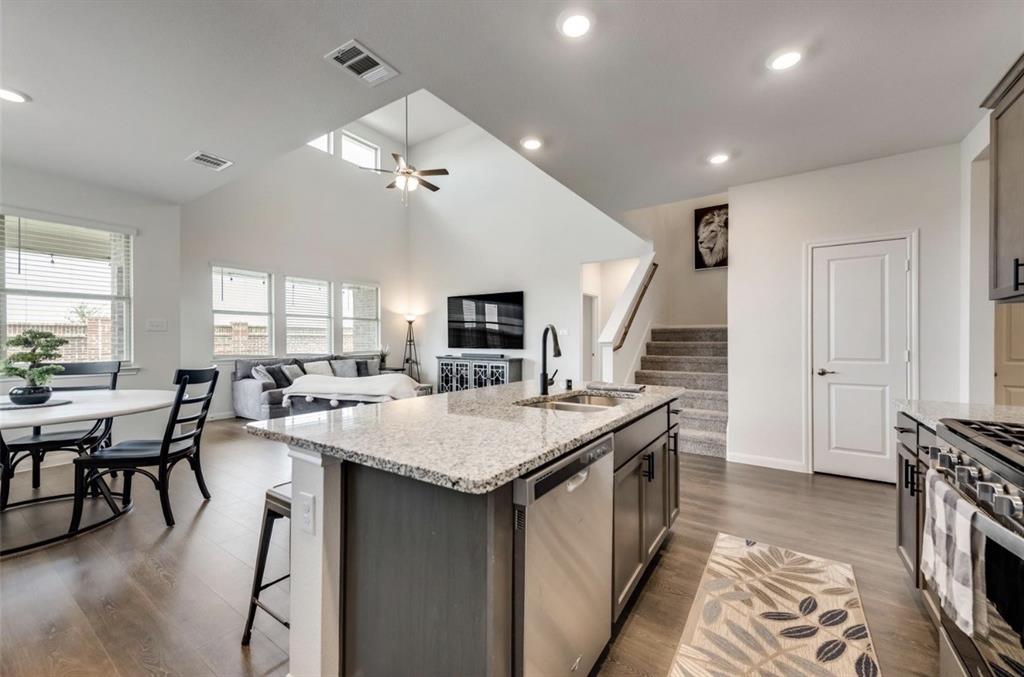 Open-concept kitchen and living area featuring a large center island with granite countertop and dark cabinetry, stainless steel appliances including a dual-fuel range, recessed and pendant lighting, vaulted ceilings with skylights, hardwood-finish flooring, and direct sightlines to a multi-level staircase and secondary living spaces