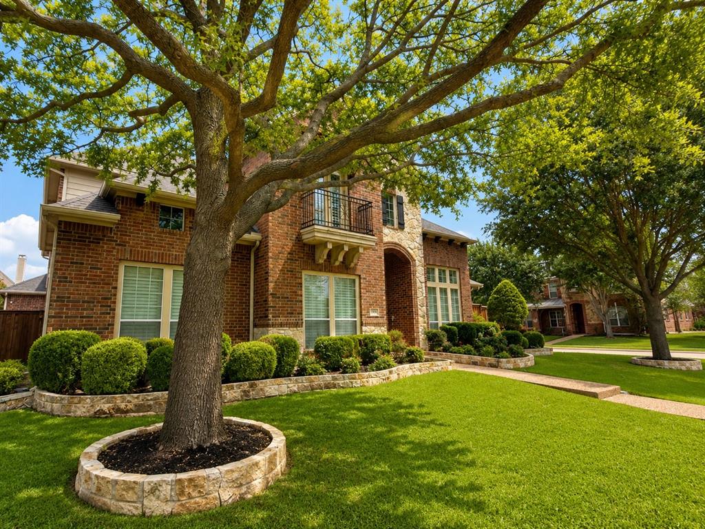 Brick and stone exterior featuring a second-story balcony with iron railing, manicured front lawn, and mature trees