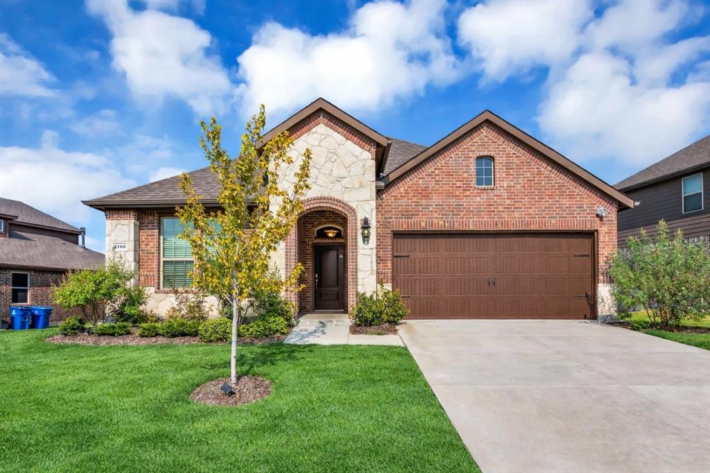 Brick and stone facade featuring a prominent arched entryway, a two-car garage with a panel door, a concrete driveway, and a landscaped front yard with a young tree Brick and stone facade featuring a prominent arched entryway, a two-car garage with a panel door, a concrete driveway, and a landscaped front yard with a young tree
