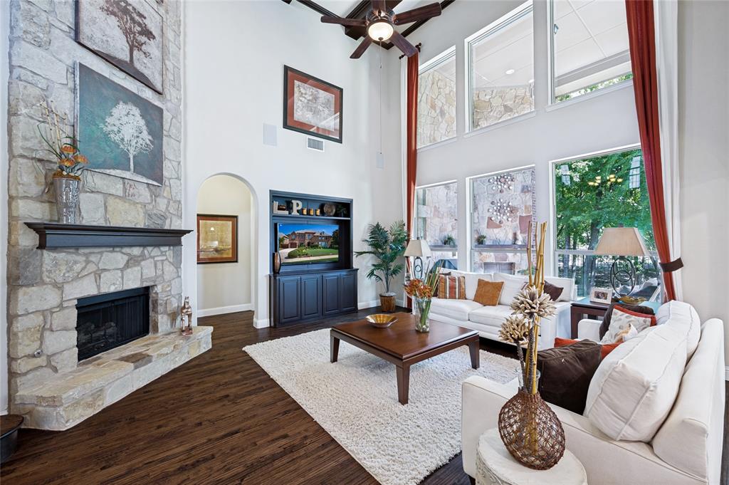 Two-story living area featuring a floor to ceiling fireplace with mantel, hardwood flooring, built-in entertainment center, and multiple large windows with view of mature trees.