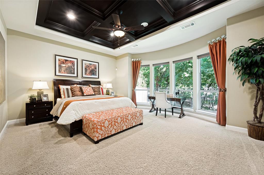 The spacious primary suite features panoramic views of the surrounding trees, a coffered ceiling with recessed lighting, a ceiling fan, a curved wall accented by multiple windows, and a generous sitting area.