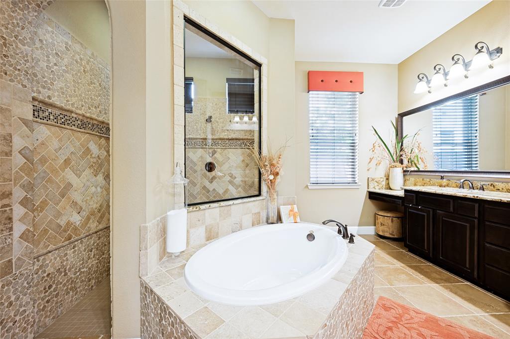 This primary bathroom features natural light, a large soaking tub with custom tile surround, a walk-in double shower accented by pebble and herringbone tile work, dual vanities including a dedicated makeup station, a linen closet, and expansive framed mirrors.