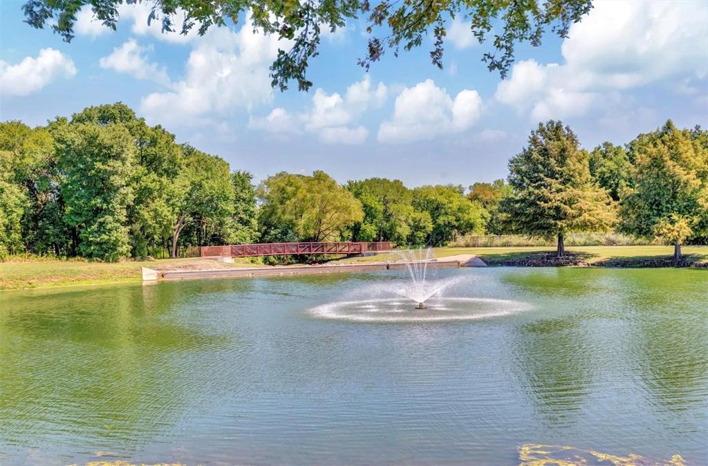 Pond with a central fountain, surrounded by mature trees, a grassy bank, and a pedestrian bridge