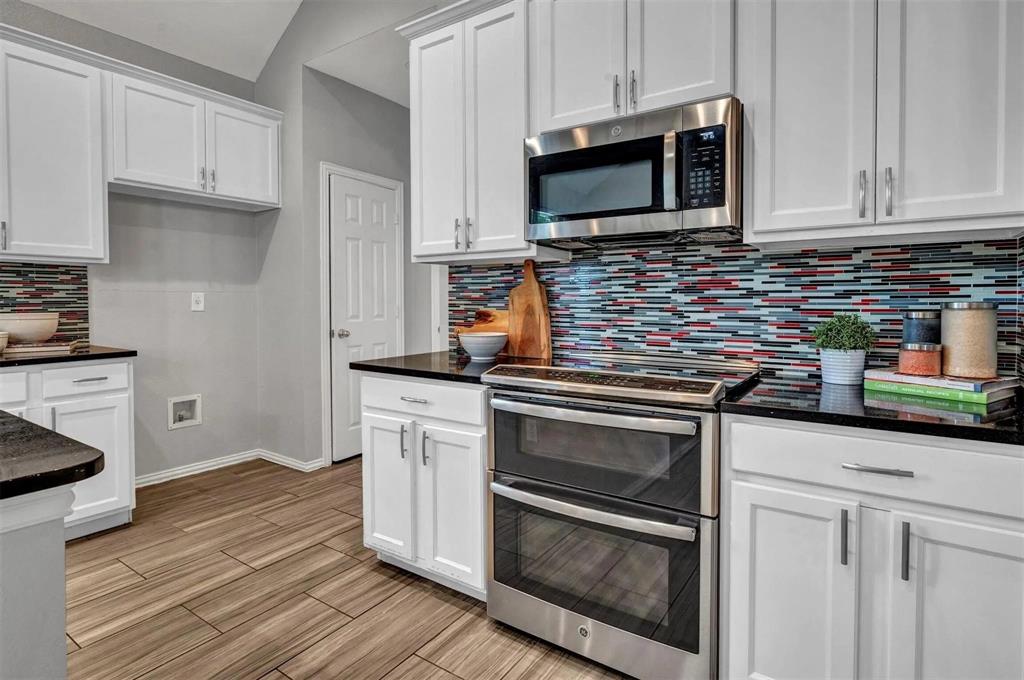 White shaker-style cabinetry, black countertops, stainless steel appliances, mosaic tile backsplash, and wood-finish flooring