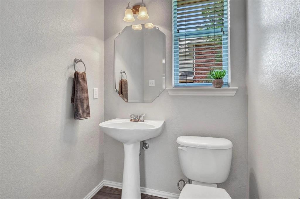 Pedestal sink, wall-mounted light fixture, decorative mirror, window with blinds, and wood-finish flooring