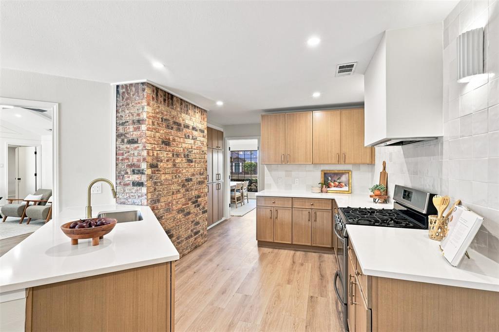 Modern kitchen featuring dual islands with wood-finish cabinetry and white quartz countertops, stainless steel gas cooktop, custom hood ventilation, exposed brick accent wall, and recessed ceiling lighting throughout the open layout with hardwood flooring
