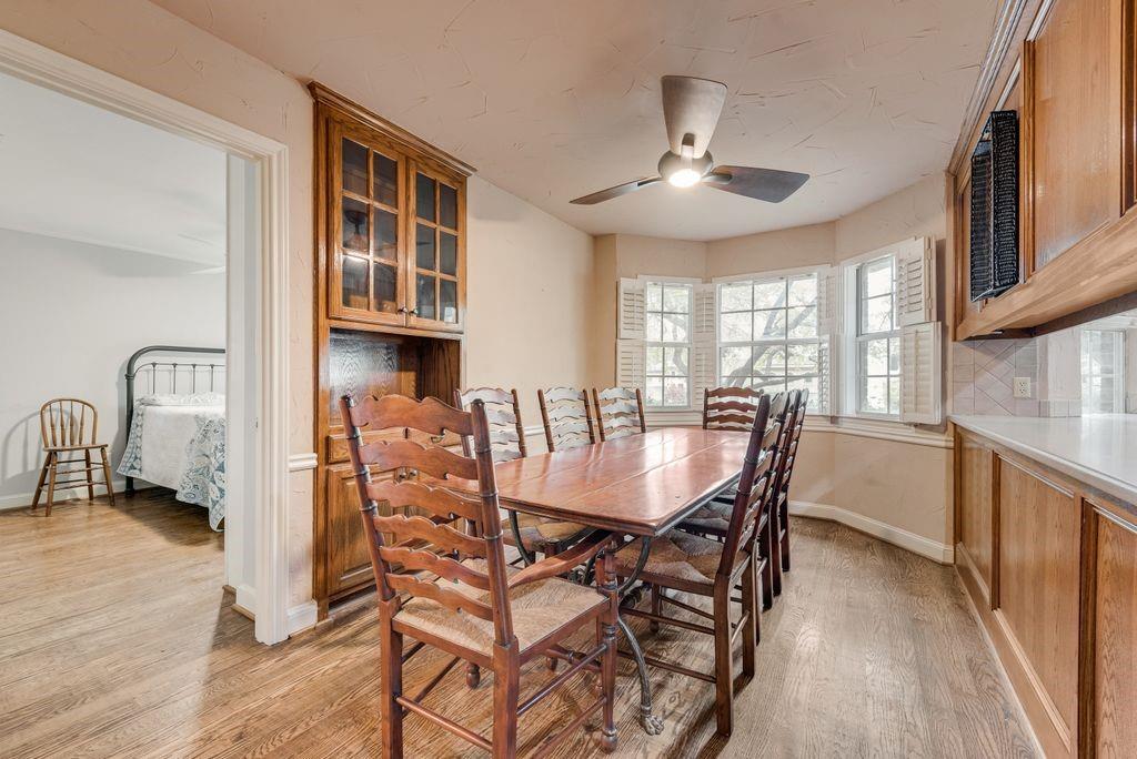 Wood-finish flooring throughout, built-in cabinetry with glass display, bay window with plantation shutters, ceiling fan with integrated lighting, and a neutral color palette