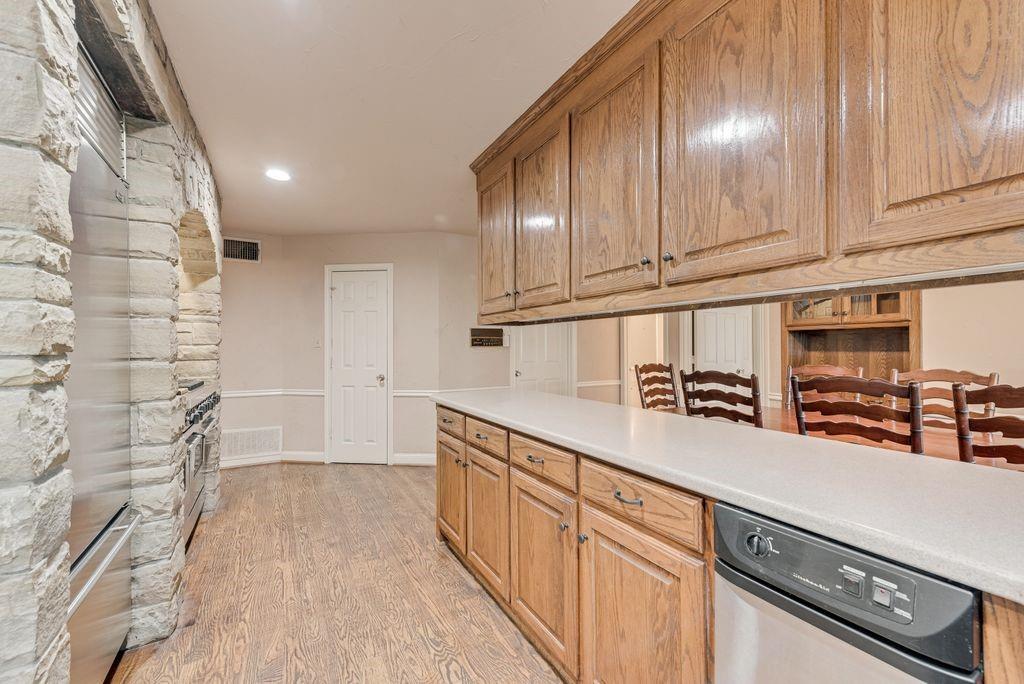 Wood-finish flooring, stone-clad archway, oak cabinetry, light-toned countertops, and a built-in stainless steel dishwasher
