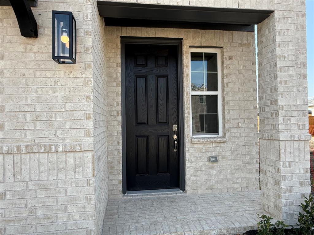 Light-toned brick exterior with a dark-paneled entry door, a single-pane window with grid inserts, an exterior wall lantern, and a covered porch Light-toned brick exterior with a dark-paneled entry door, a single-pane window with grid inserts, an exterior wall lantern, and a covered porch