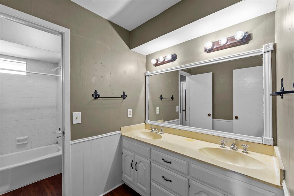 Dual vanity with integrated sinks and white cabinetry, large framed mirror, wood-finish flooring, and a tiled tub shower