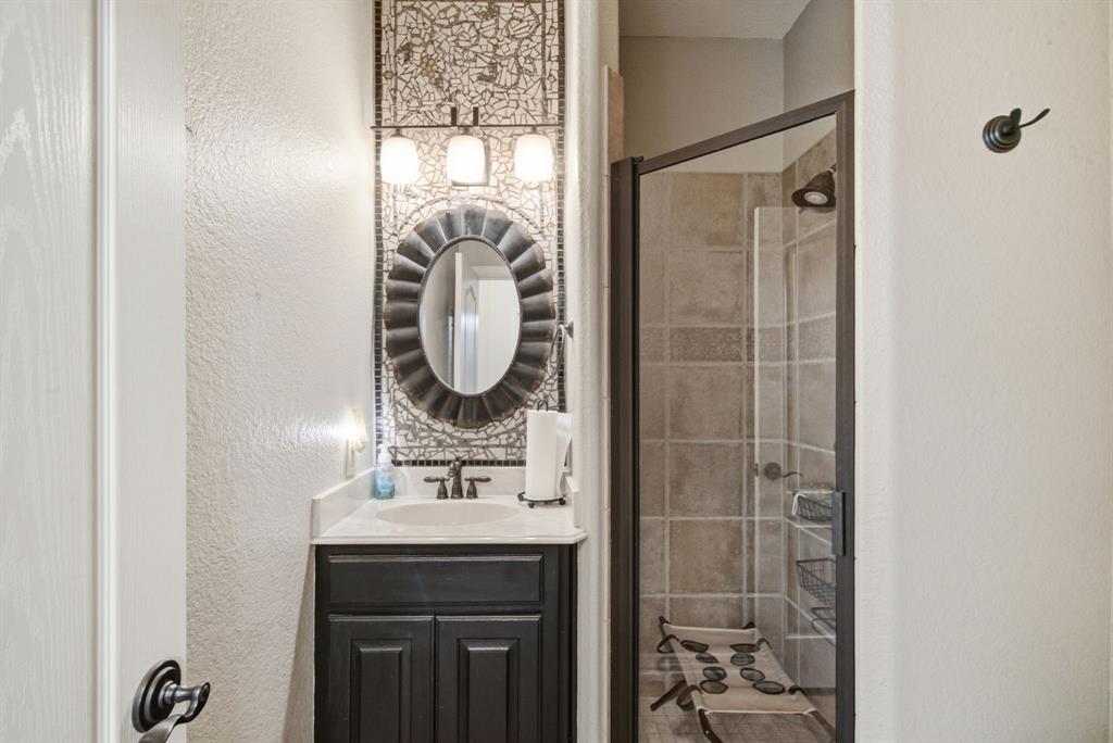 Downstairs bathroom featuring a dark-finish vanity with a light-toned countertop, a decorative tiled wall accent, a scalloped-edge mirror, and a tiled shower enclosure with a dark-finish frame