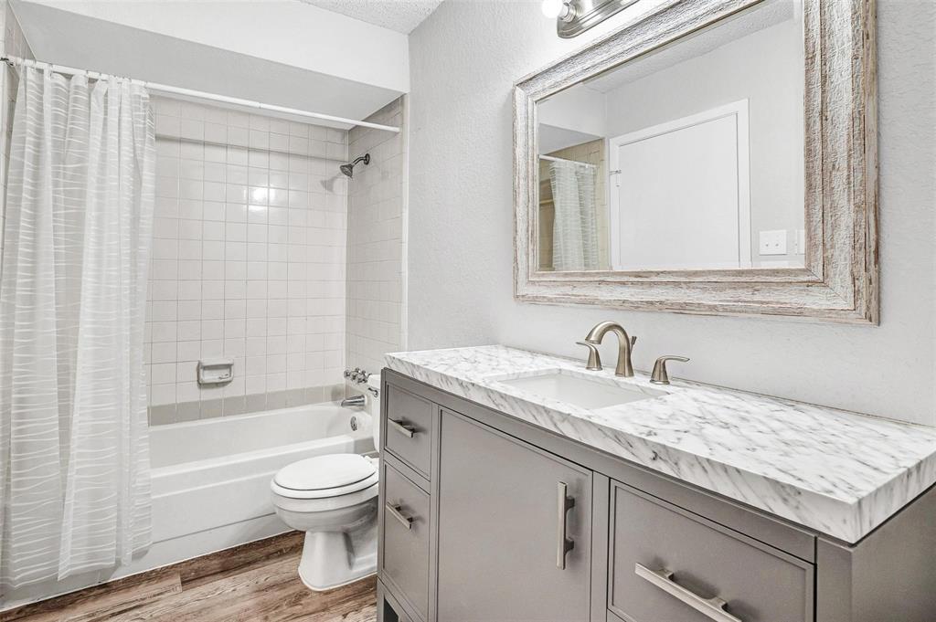 Vanity with a white countertop and integrated sink, framed mirror, built-in bathtub with white tile surround, and wood-finish flooring