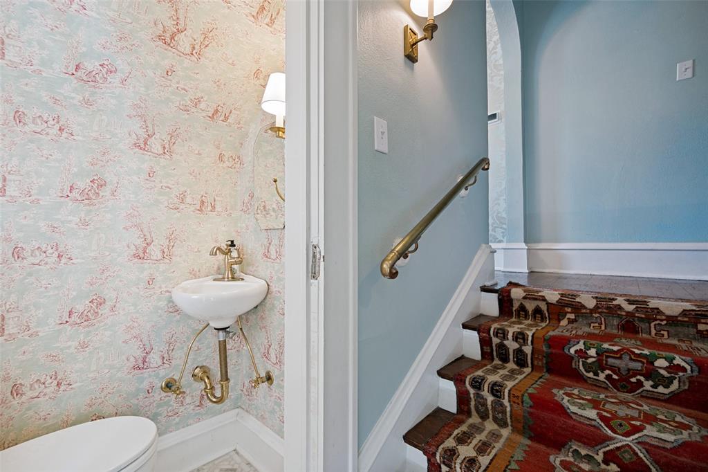Pedestal sink with brass-finish fixtures, patterned wallpaper, and a staircase with a brass handrail