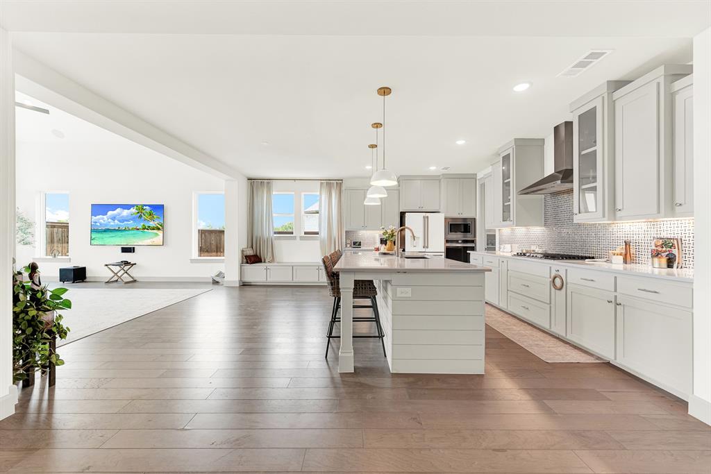 A long sightline spans across warm hardwood floors into a sunny wall of windows. It’s a welcoming spot where kids can finish homework at the counter while dinner simmers.