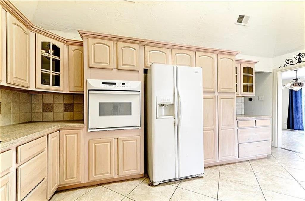 Spacious kitchen featuring light wood cabinetry with raised-panel doors, ceramic tile backsplash, white side-by-side refrigerator, white built-in wall oven with microwave, and neutral tile flooring throughout the cooking and dining area