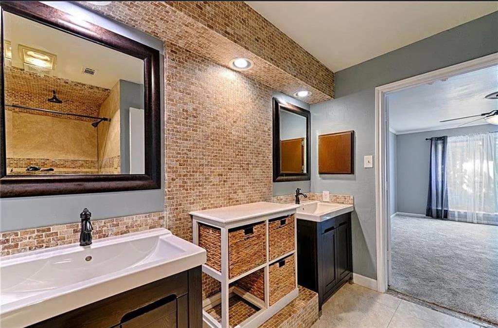 Master bathroom featuring dual vanities with wood-finish cabinetry and rectangular vessel sinks, brick accent wall with recessed lighting, large framed mirror, and tile flooring throughout