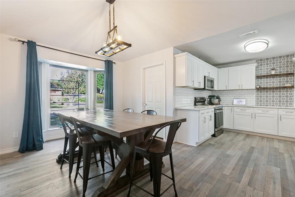 Open-concept kitchen and dining area featuring white cabinetry with stainless steel appliances, patterned tile backsplash, wood-finish flooring throughout, and large windows with blue drapery overlooking landscaped grounds Open-concept kitchen and dining area featuring white cabinetry with stainless steel appliances, patterned tile backsplash, wood-finish flooring throughout, and large windows with blue drapery overlooking landscaped grounds