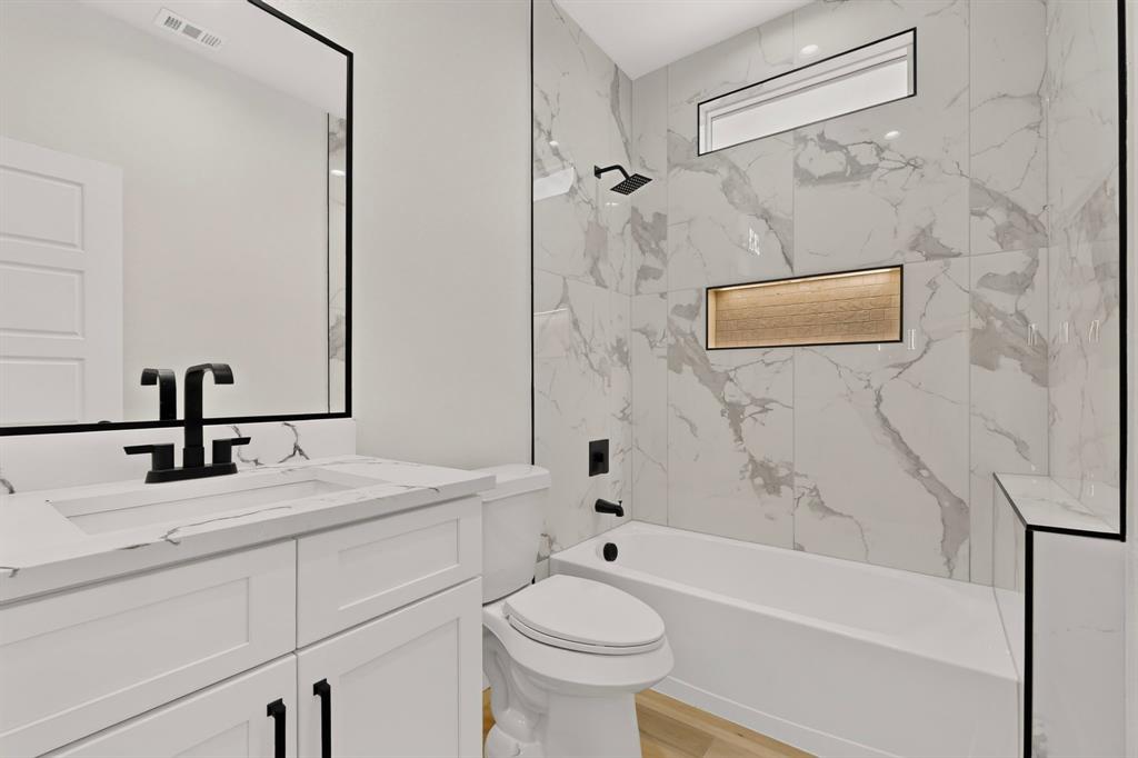 White vanity with integrated sink and matte black faucet, framed mirror, full-height wall tiling, built-in bathtub, and wall niche with textured accent