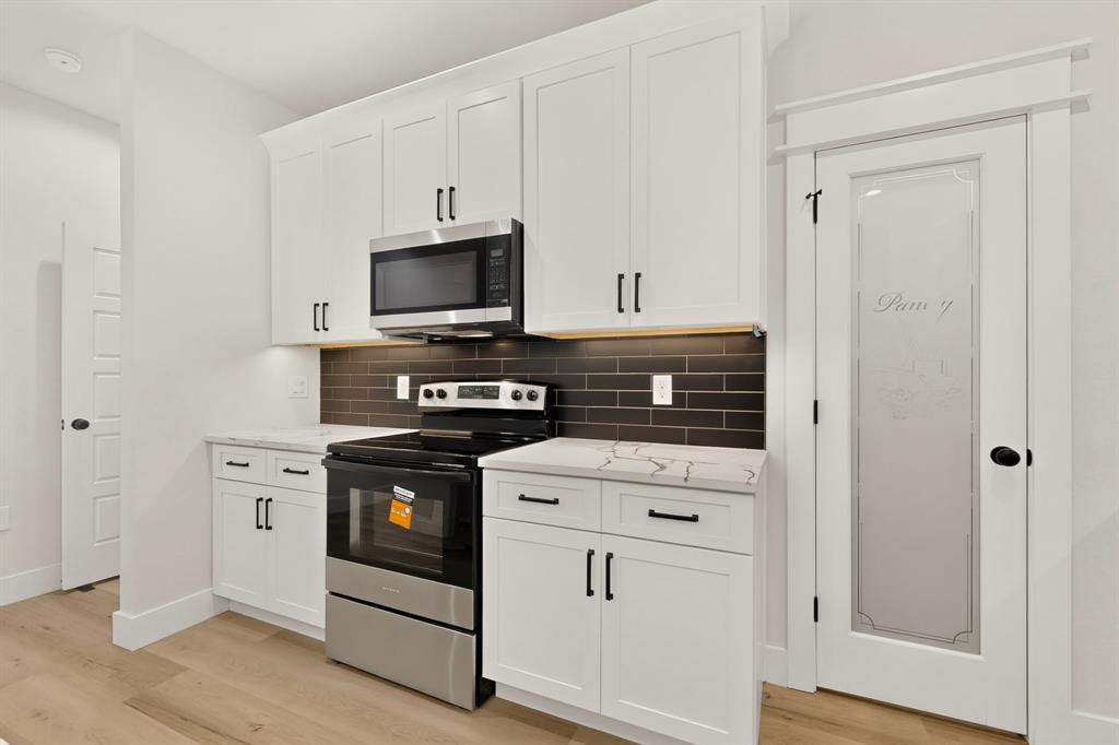 White shaker cabinetry with black hardware, stainless steel appliances, dark subway tile backsplash, light wood-finish flooring, and a frosted glass pantry door