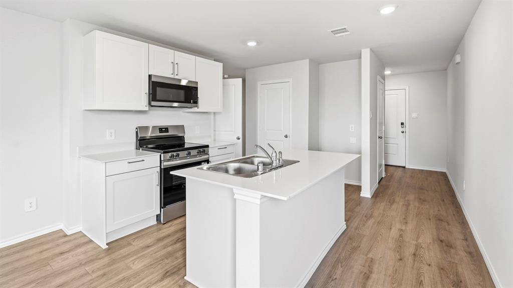 Kitchen featuring white cabinetry, stainless steel appliances, a kitchen island with an integrated sink, white countertops, and wood-finish flooring Kitchen featuring white cabinetry, stainless steel appliances, a kitchen island with an integrated sink, white countertops, and wood-finish flooring