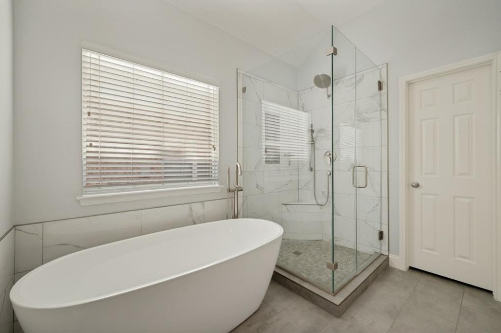 Freestanding soaking tub with floor-mounted faucet, glass-enclosed shower with tile surround and bench, and large format tile flooring