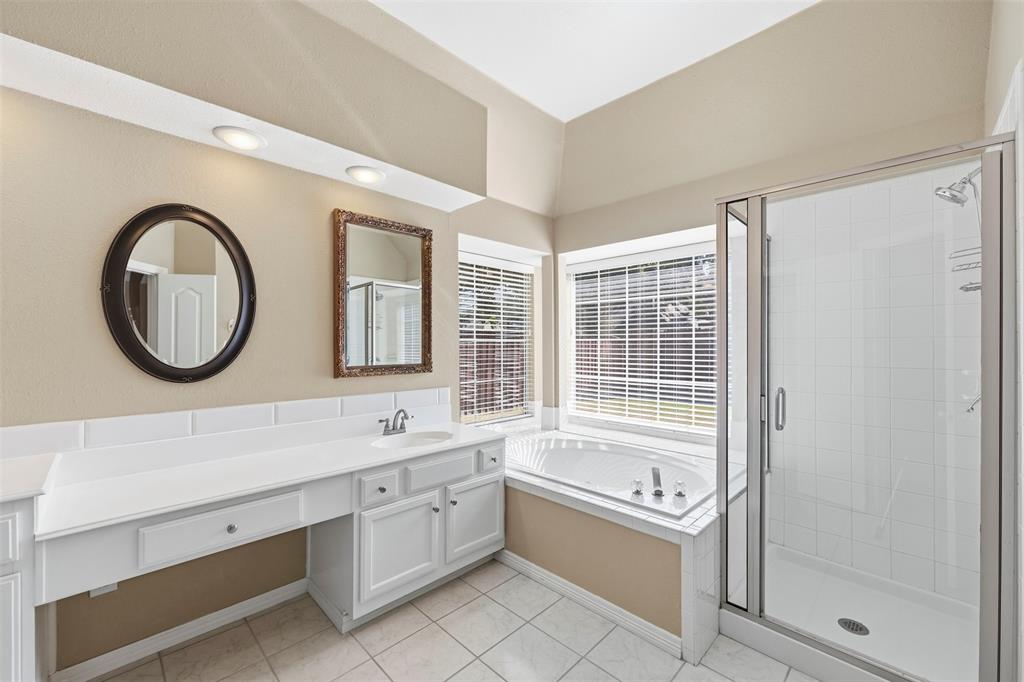 White vanity with integrated sink, expansive countertop, white tile flooring, built-in corner bathtub, and a glass-enclosed shower