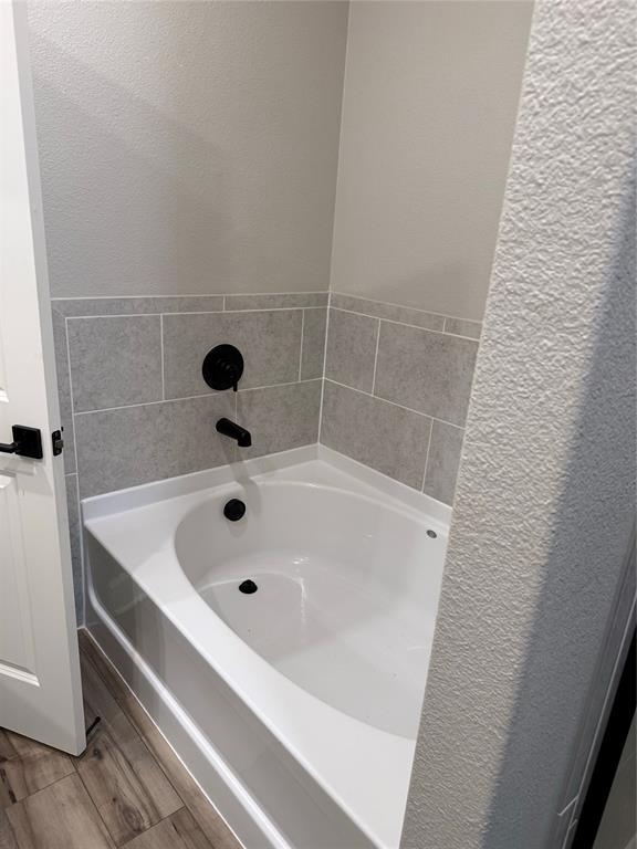White bathtub with matte black fixtures, grey tiled surround, and wood-finish flooring